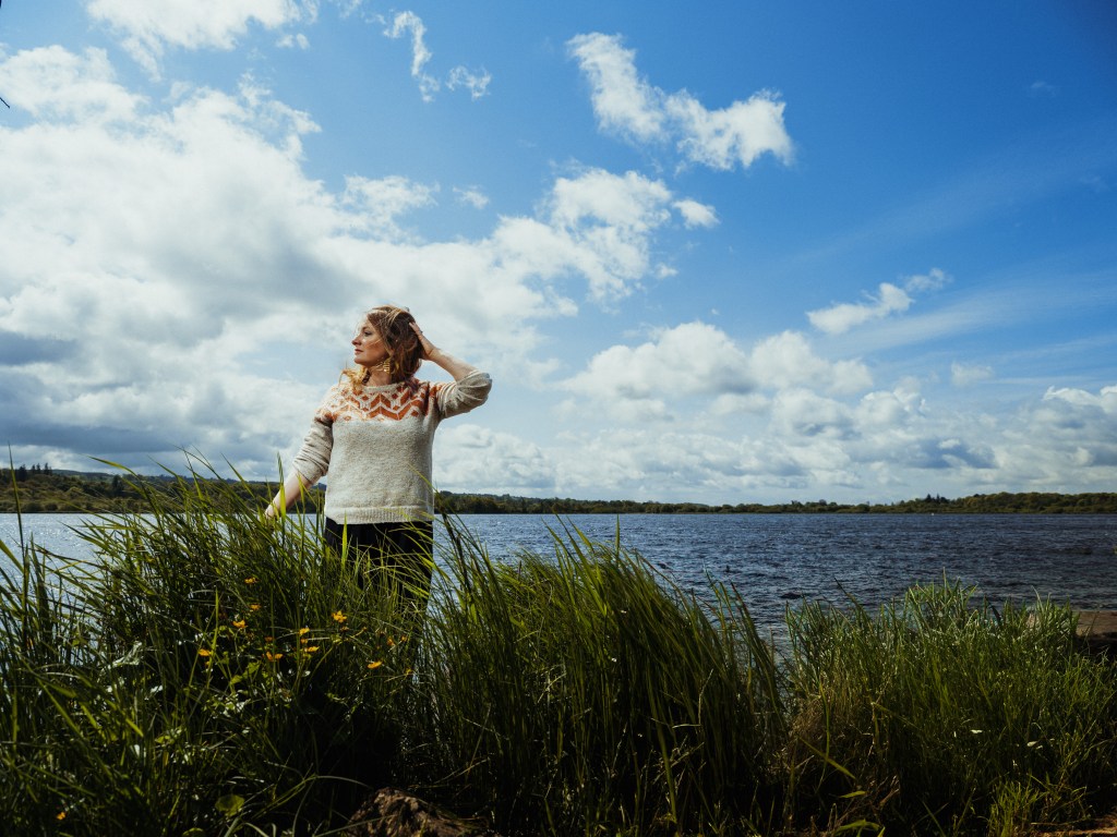 A woman in a cosy jumper and big earrings stands in front of a loch and a big blue sky. In the foreground there is long grass. She's holding her hair back in the wind.